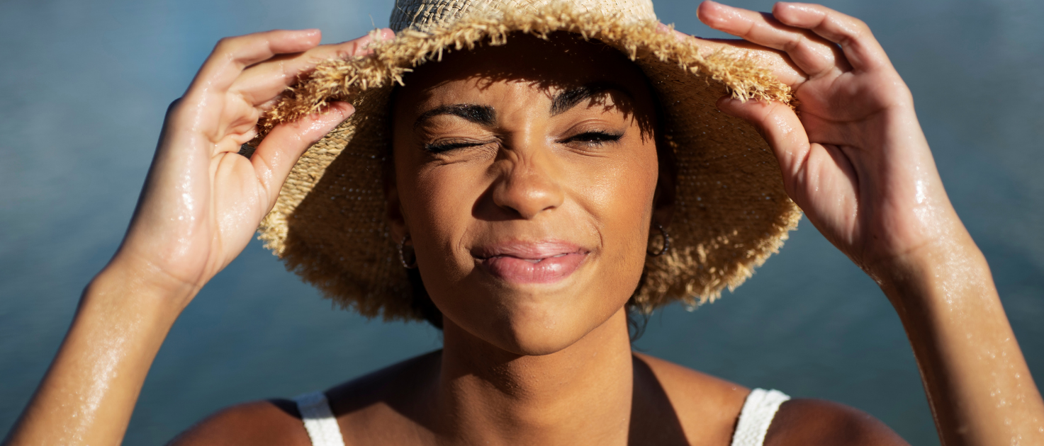 Mulher ao ar livre usando chapéu de palha, segurando a aba com as mãos, iluminada pela luz do sol, com água ao fundo.