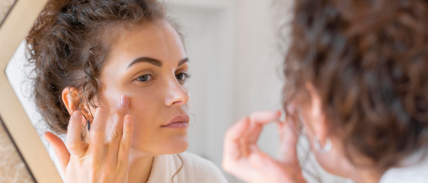 Mulher aplicando creme no rosto em frente ao espelho, cuidando da pele de forma preventiva.